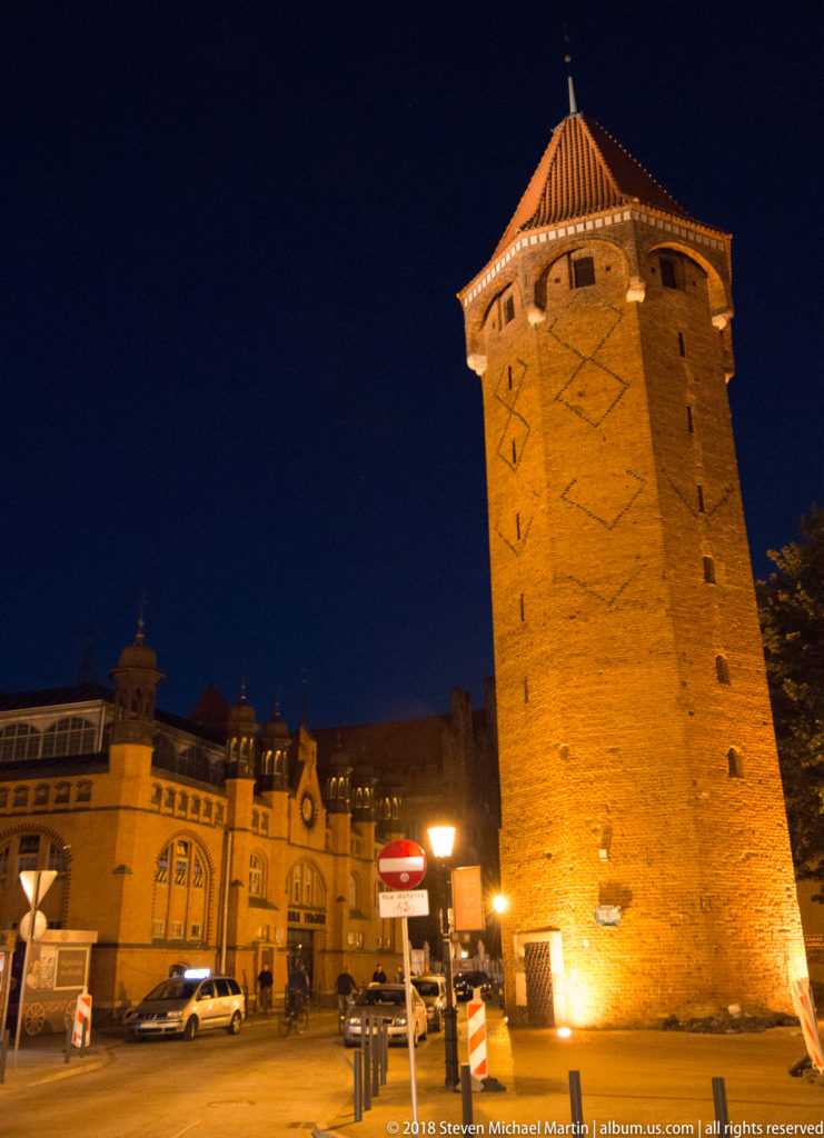 Floodlit square brick Jacek Tower with steep tiled roof and small openings, beside part of Hala Targowa.