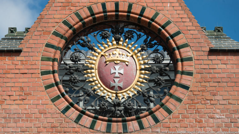 Crowned Gdańsk coat of arms with two crosses in a black metal grille and circular brick opening.