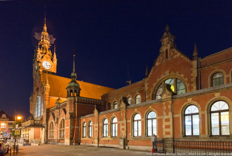 Illuminated red-brick Gdańsk Główny station with large arched windows, shaped gables, steep roof, and ornate clock tower.