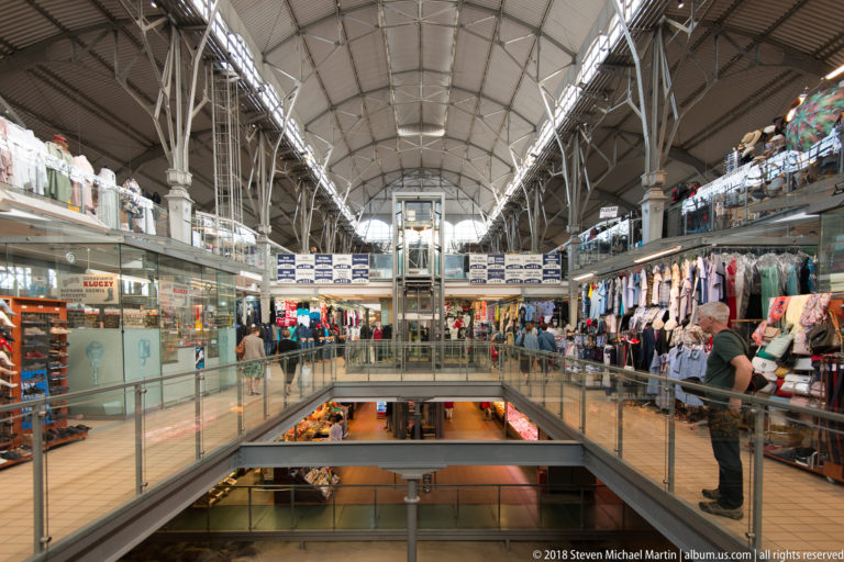 Multi-level covered market with clothing stalls, glass railings, central elevator and high metal-truss roof.
