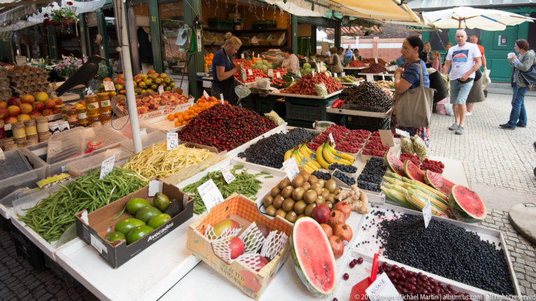 Outdoor market stall piled with berries, melons, beans, fruit and vegetables as shoppers browse.
