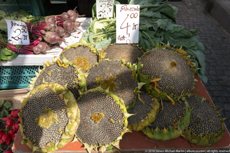 Large seed-filled sunflower heads stacked on a market table beside leafy vegetables and handwritten price signs.