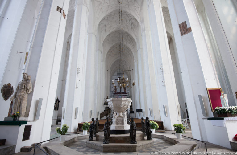 White vaulted nave of St. Mary’s Basilica with large stone baptismal font and dark sculptural figures in foreground.