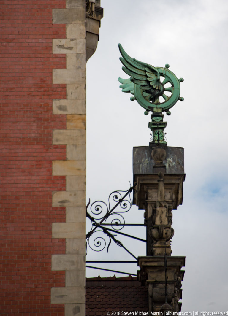 Green-patinated winged metal emblem on a stone pier beside red brick and wrought-metal scrollwork.