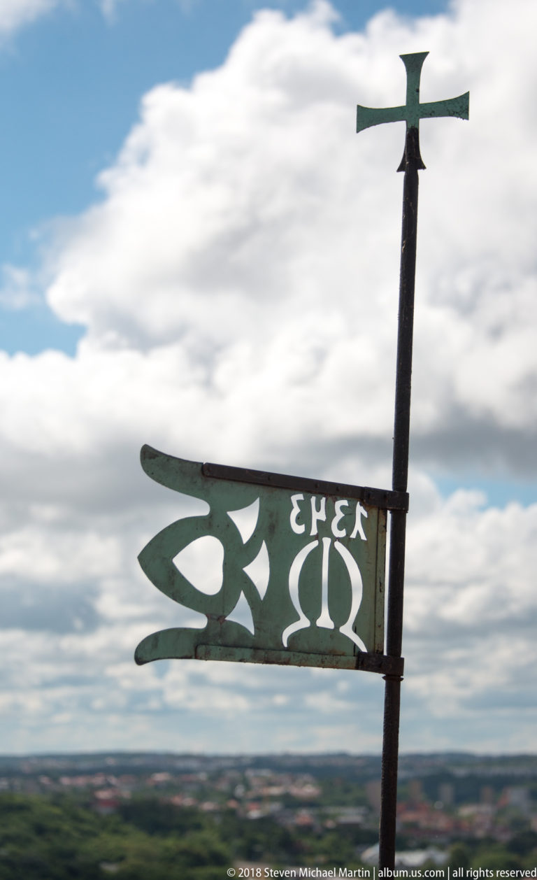 Openwork metal flag-shaped weather vane topped by a cross, with distant landscape blurred below.