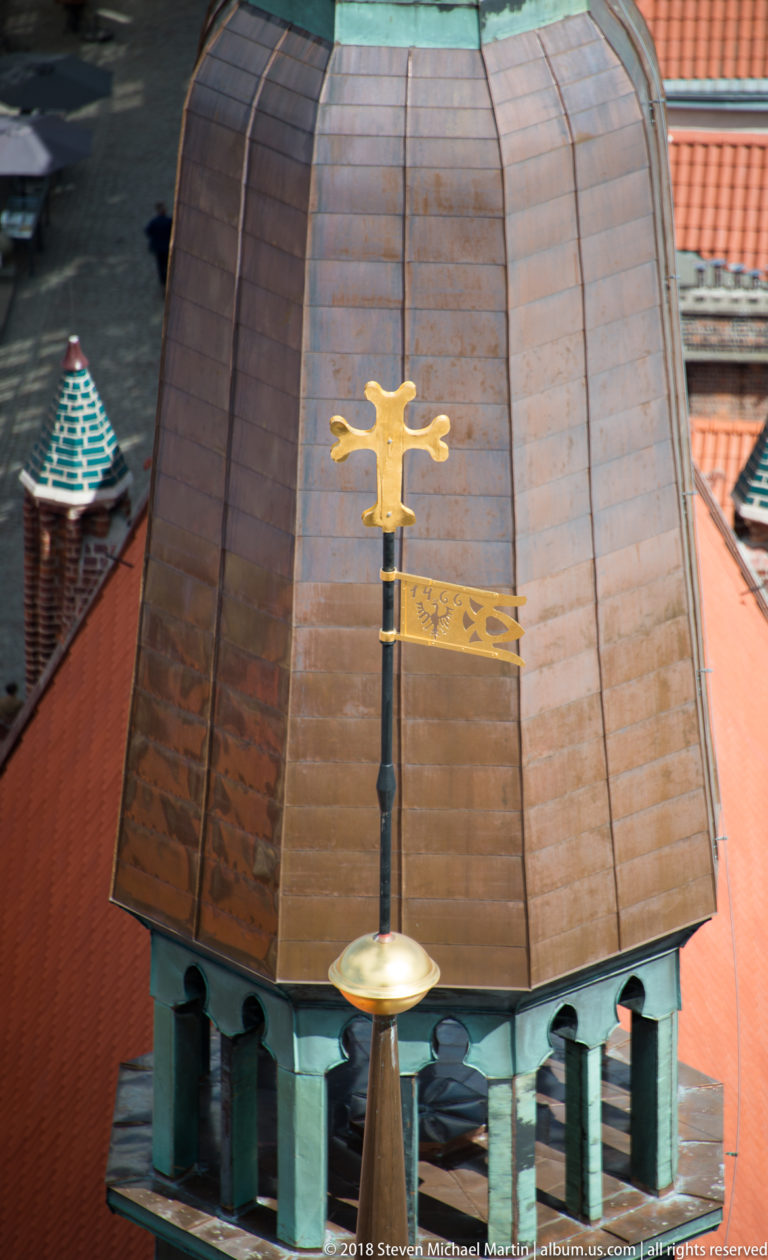 Gilded flag-shaped weather vane in front of a broad weathered copper roof and surrounding red tiles.