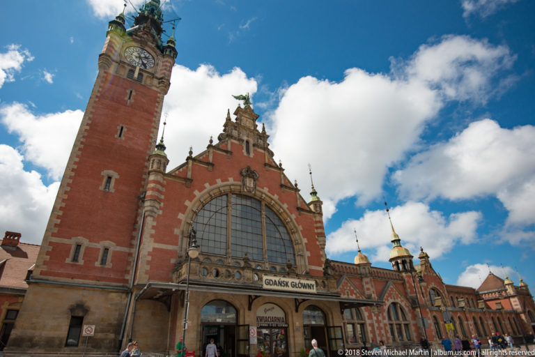 Low-angle view of Gdańsk Główny station with tall brick clock tower, arched entrance window and pedestrians below.