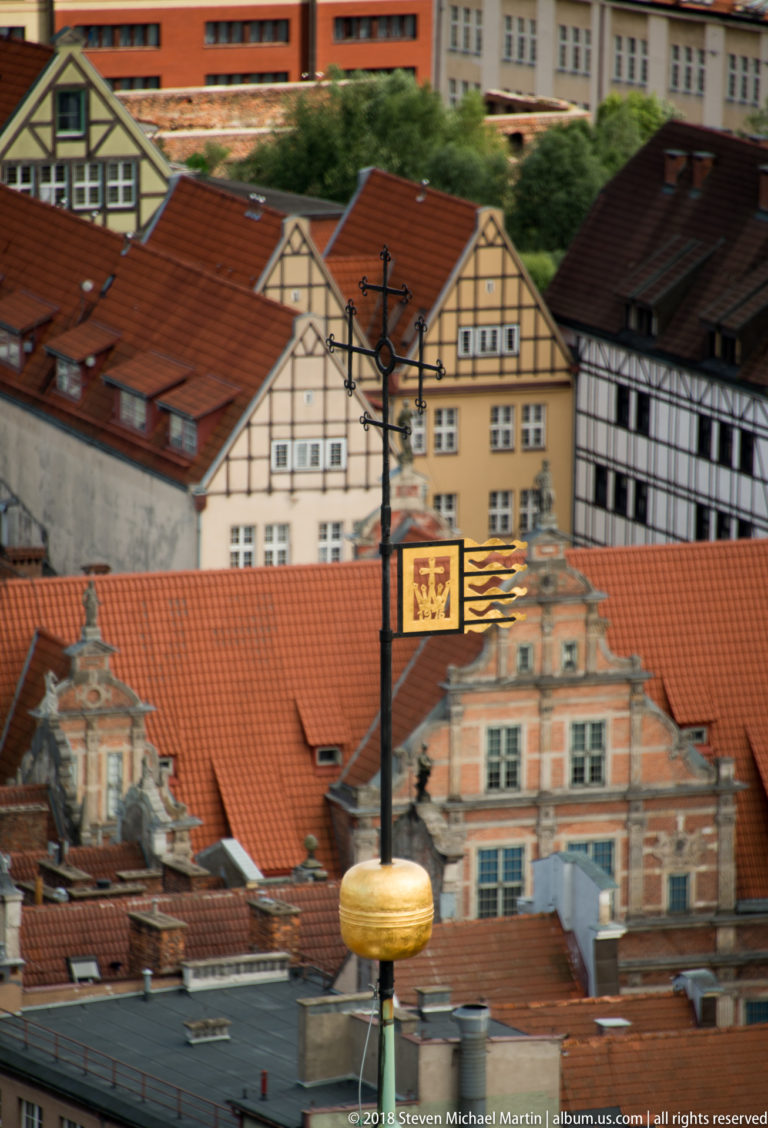 Red-tile rooftops and colorful historic façades behind an ornate metal bracket with a gilded hanging oval sign.