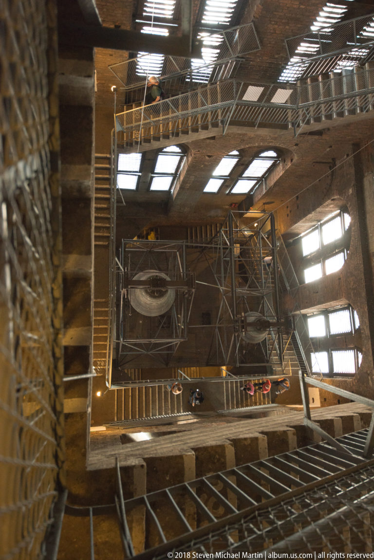 Exposed timber roof trusses, braces, skylights, and an elevated walkway inside a large historic building.