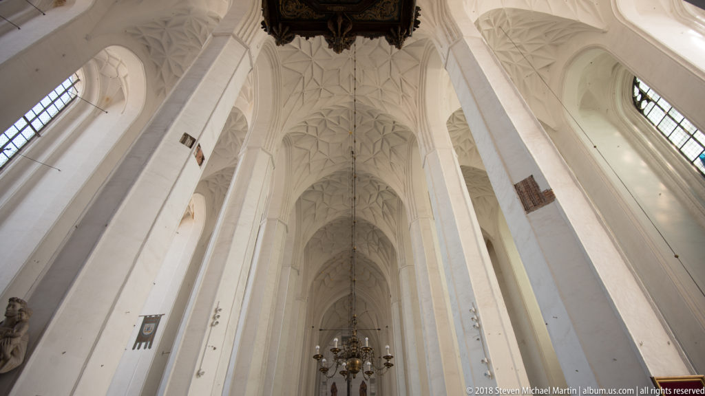 Upward view of tall pale church piers, pointed arches, high windows, and intricate Gothic vaulting.