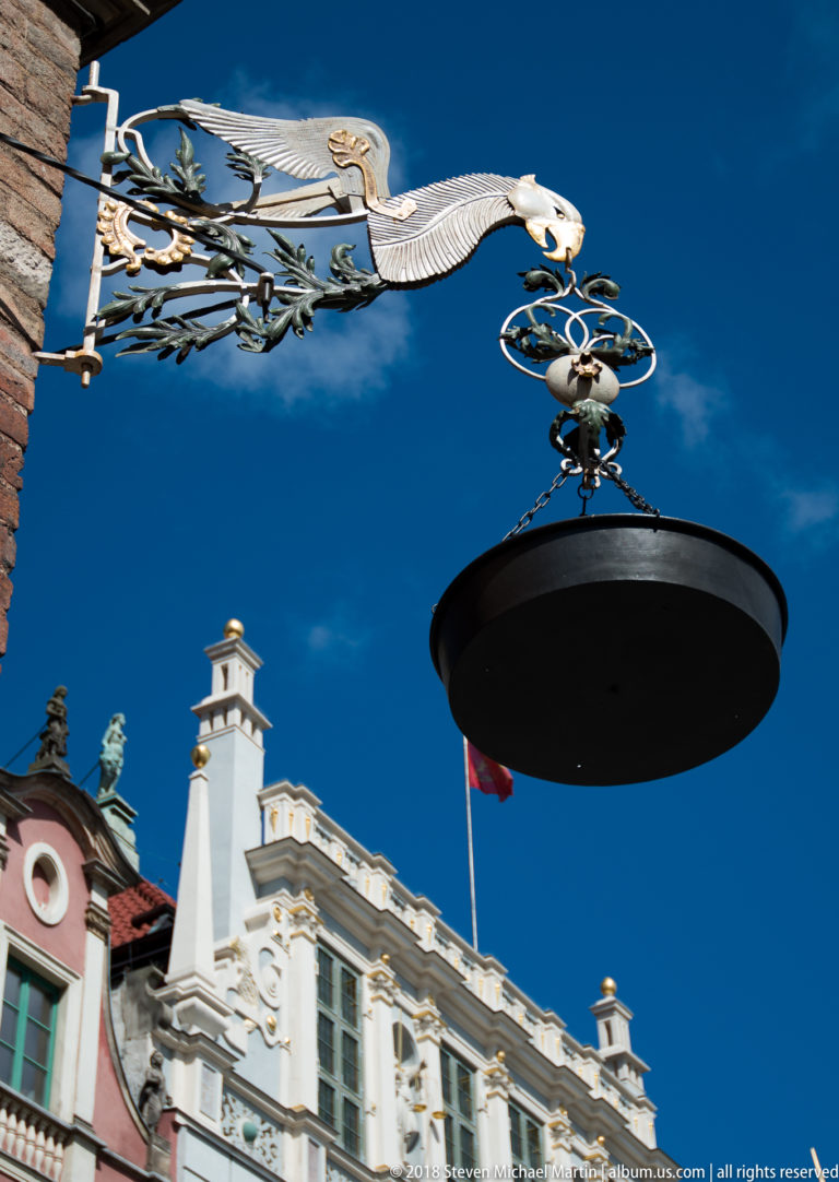 Gilded bird-shaped metal bracket supporting a black oval hanging sign above ornate historic gables.