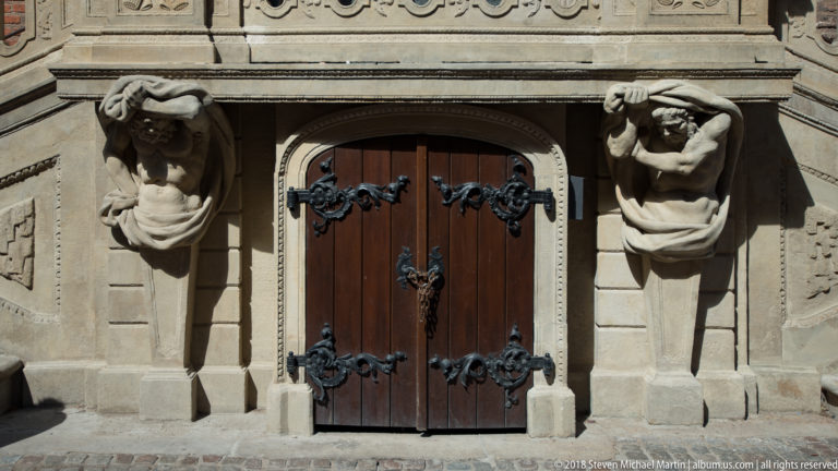 Wooden double door with black iron fittings flanked by two carved male atlantes on a stone façade.