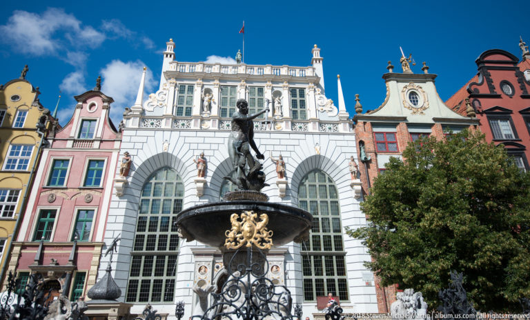Neptune Fountain in front of the white Artus Court façade and colorful gabled houses under a blue sky.