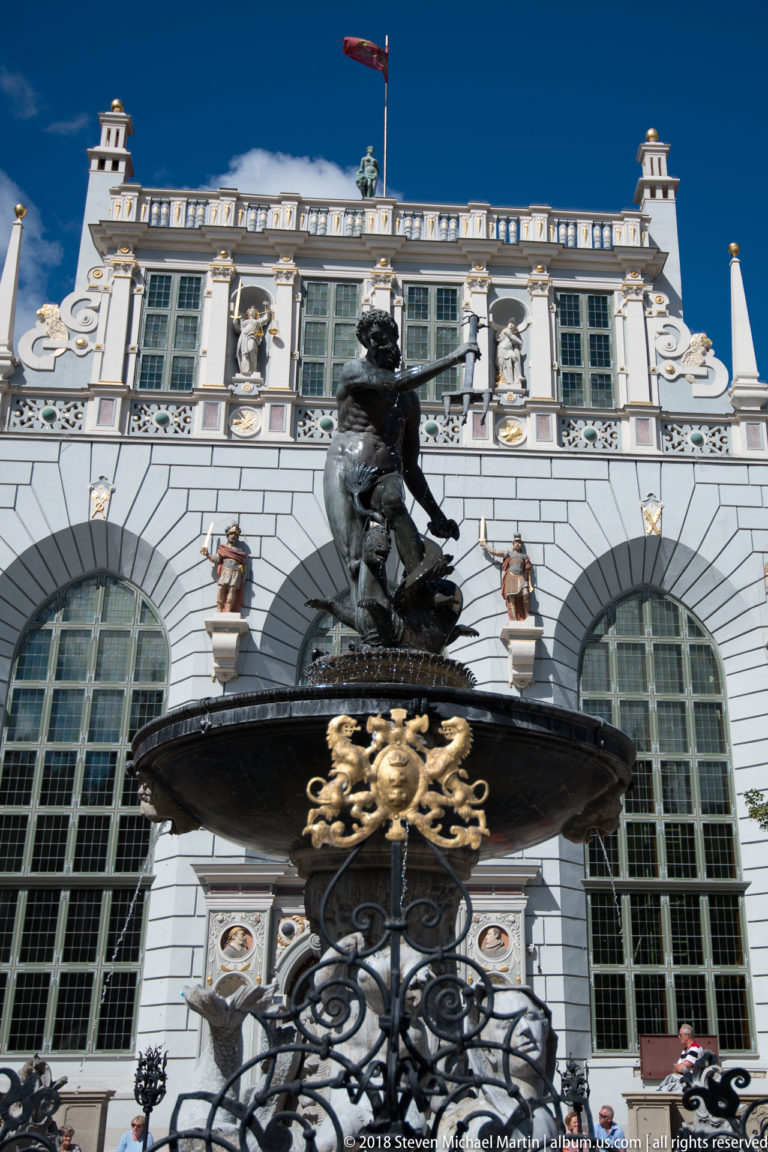 Closer view of bronze Neptune Fountain before the arched windows and sculpted white façade of Artus Court.