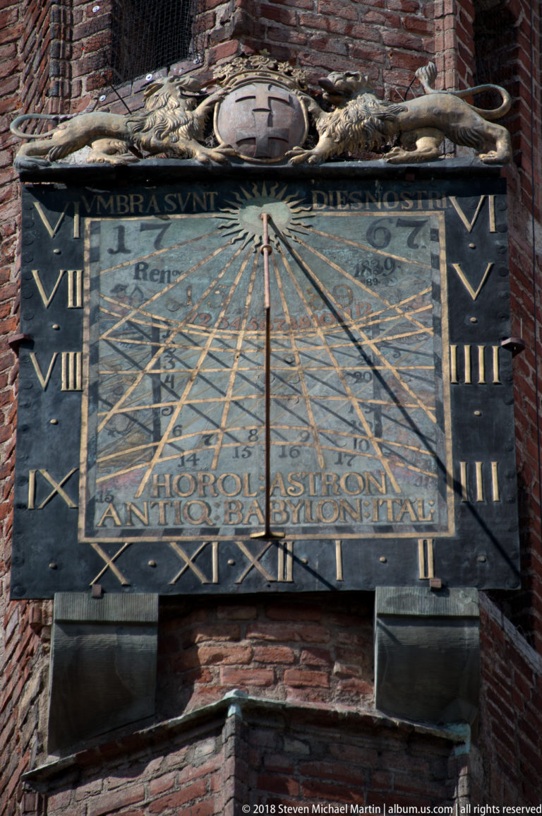 Rectangular astronomical sundial with Roman numerals, radiating lines, inscriptions, and metal gnomon on a brick wall.