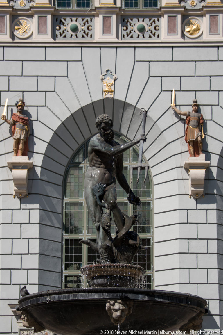 Bronze Neptune with trident before an arched Artus Court window, flanked by small painted façade sculptures.