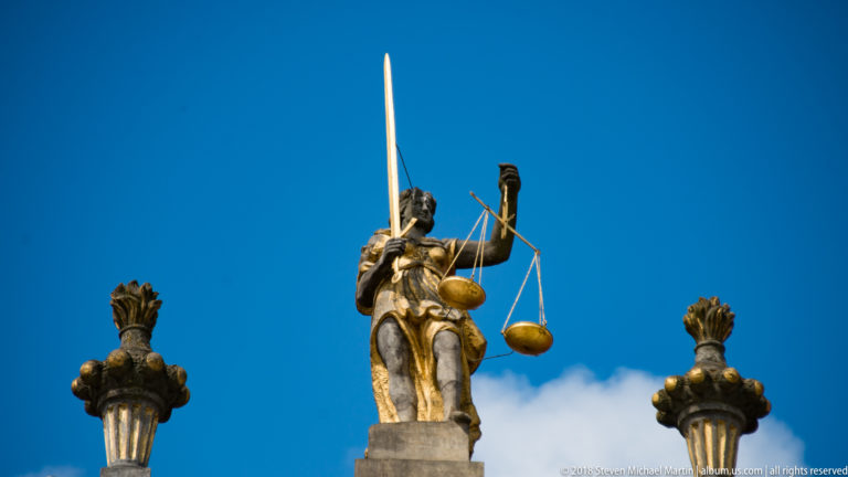 Gilded female figure of Justice holding a sword and balance scales between decorative rooftop finials.