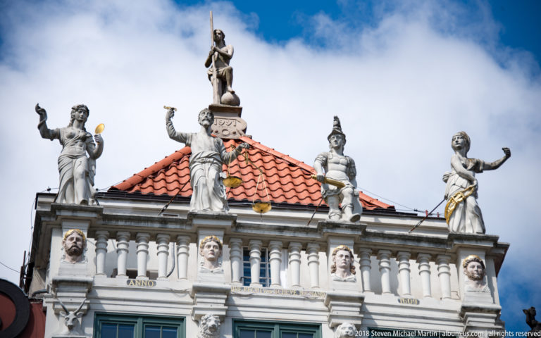 Several allegorical stone figures standing above a white balustrade and red-tile roof on an ornate historic façade.