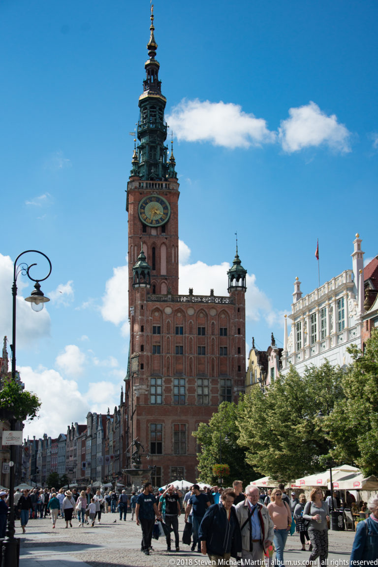 Main Town Hall tower rising above crowds, café umbrellas, trees, and historic façades along Długi Targ.
