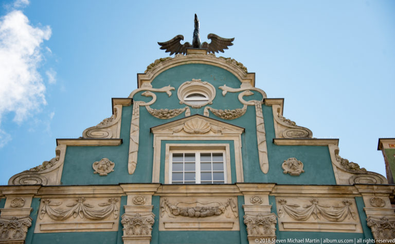 Turquoise curving gable with cream stucco scrollwork, floral reliefs, central window, and eagle finial.