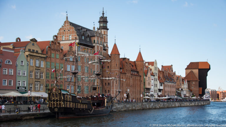 Motława River waterfront with colorful gabled houses, a historic-style tour vessel, and the Crane in the distance.