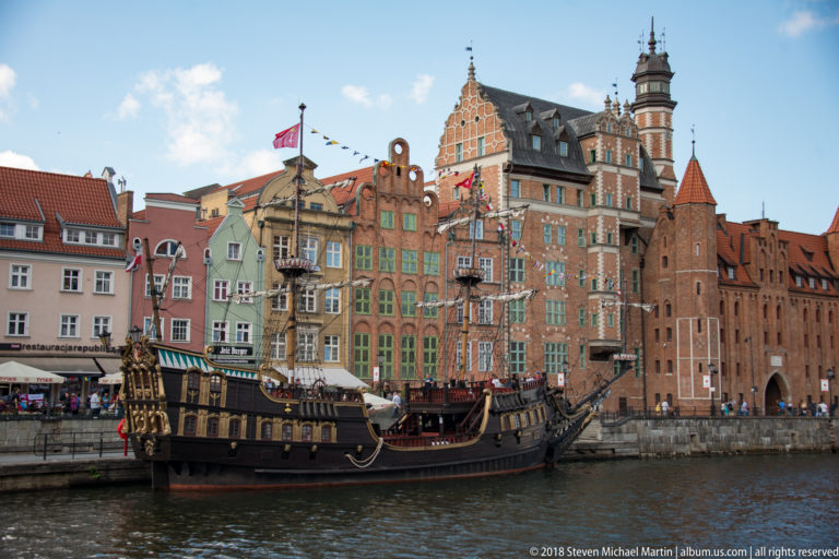 Dark historic-style tour ship moored on the Motława River before tall colorful gabled waterfront buildings.
