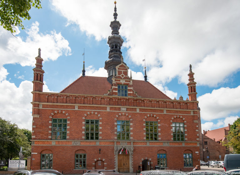 Symmetrical red-brick historic building with tiled roof, tall windows, pinnacles and a central spire.