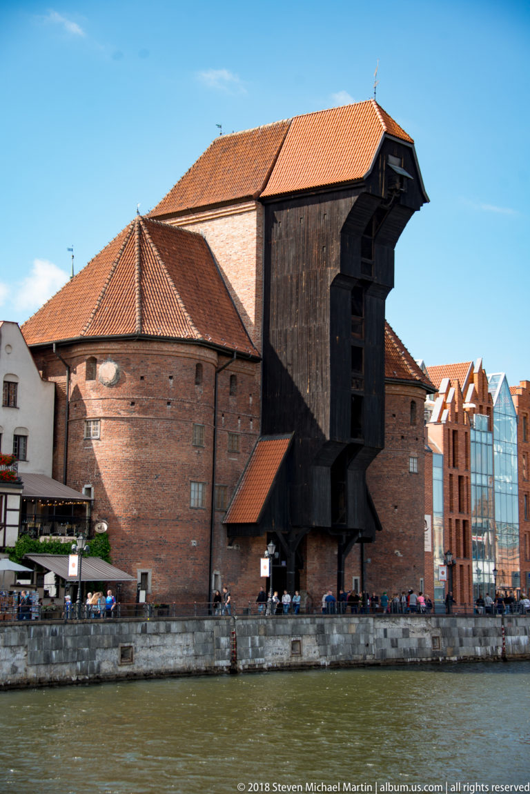 Gdańsk Crane with brick walls, red-tile roofs, and massive dark timber hoisting structure beside the Motława River.