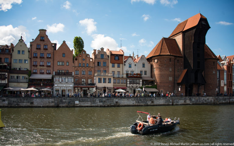 Gdańsk Crane beside gabled waterfront houses as a small motorboat crosses the Motława River in front.