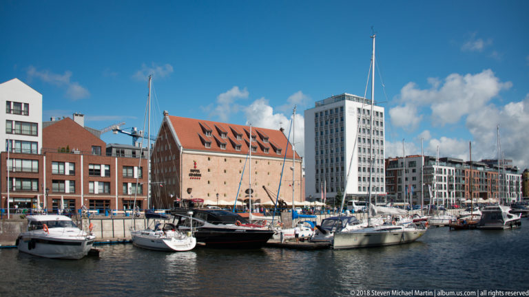 Moored sailboats and motorboats in a Motława marina backed by brick warehouses and modern waterfront buildings.