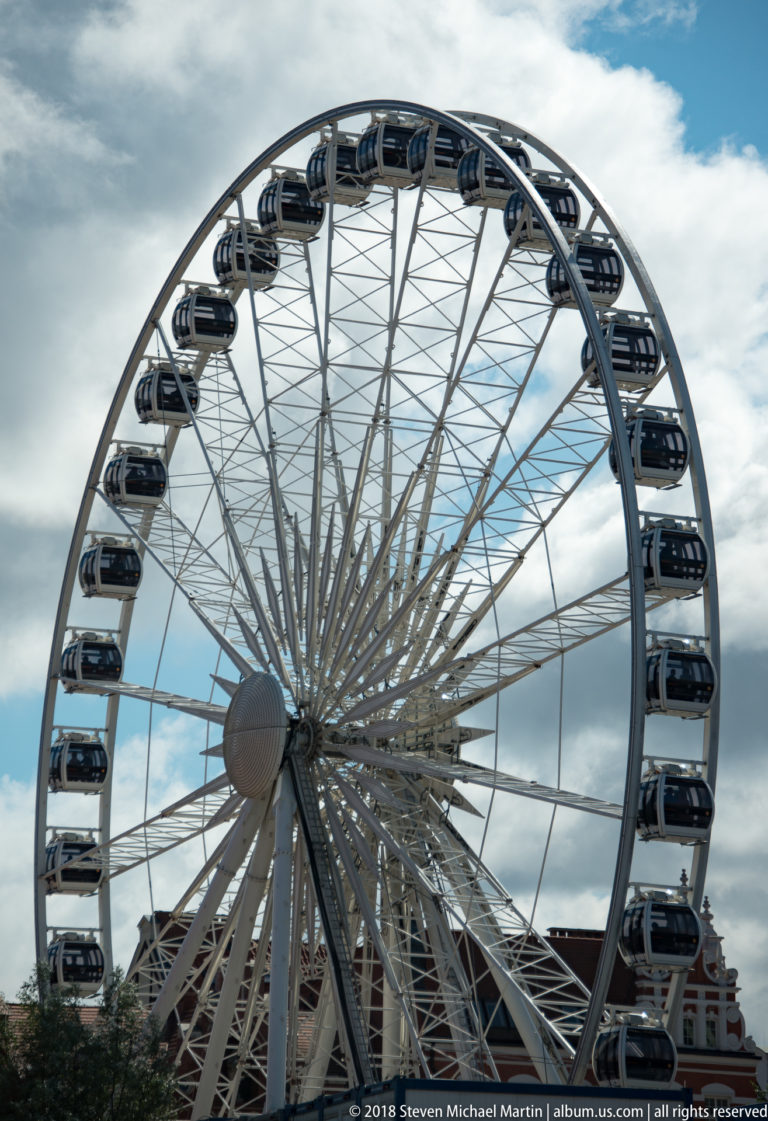 Large white observation wheel with enclosed gondolas and radiating spokes against a cloudy blue sky.