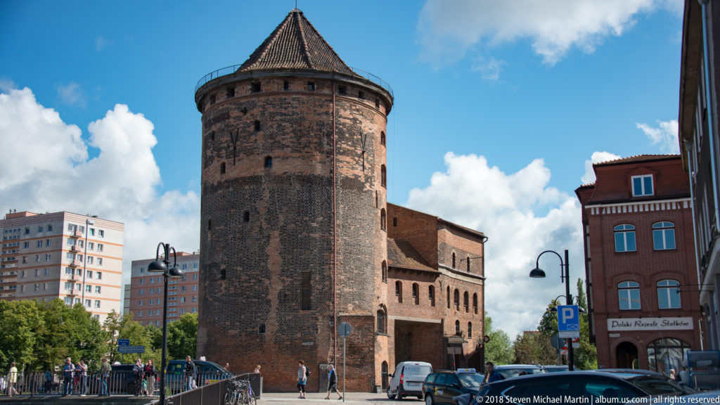 Large round medieval brick tower with conical tiled roof beside a street, with modern apartment blocks behind.