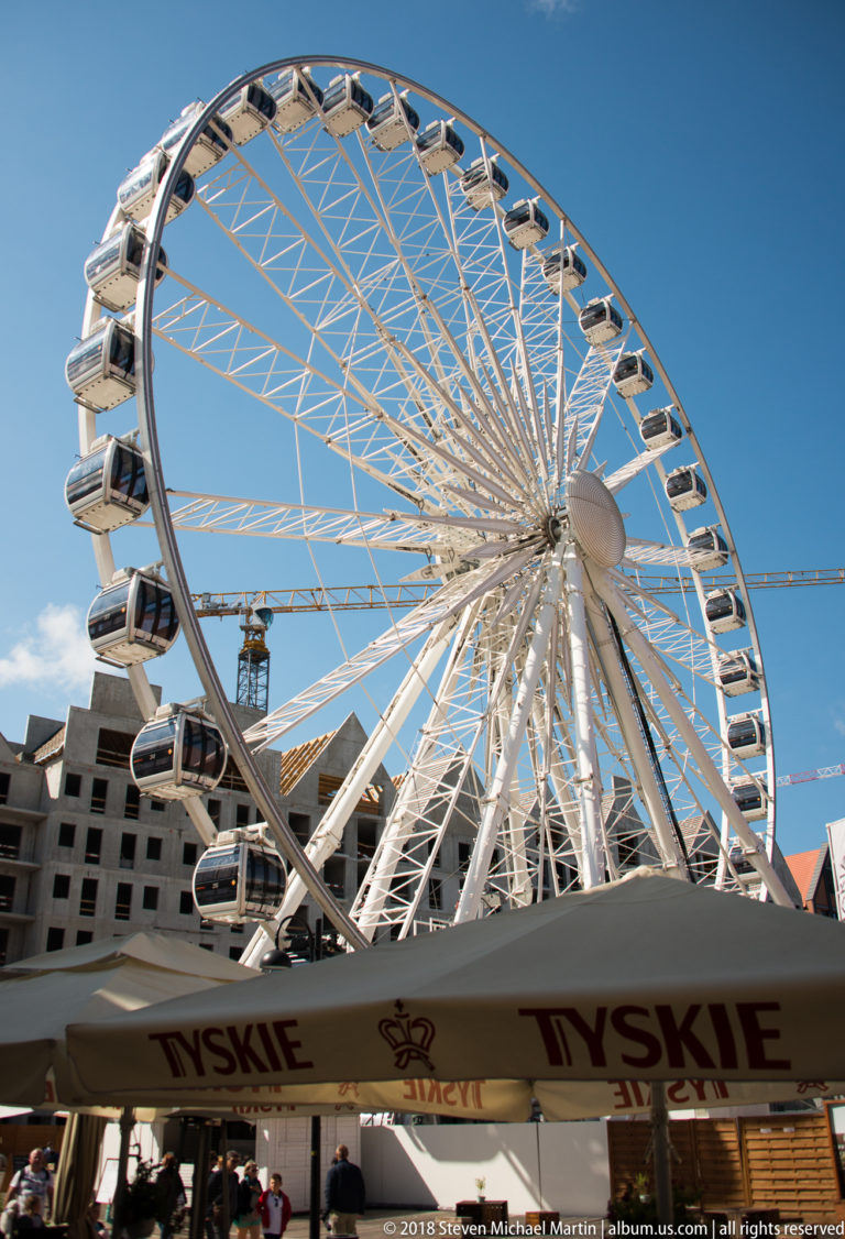 White observation wheel with enclosed gondolas rising behind café umbrellas under a clear blue sky.