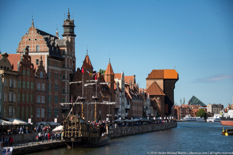 Long Motława River view with gabled waterfront buildings, a historic-style vessel, and the Gdańsk Crane downstream.