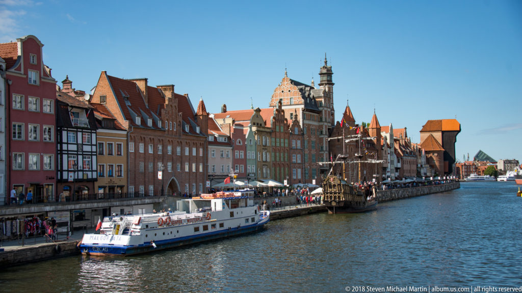 White passenger boat on the Motława beside gabled waterfront houses, a dark tour vessel, and the distant Crane.