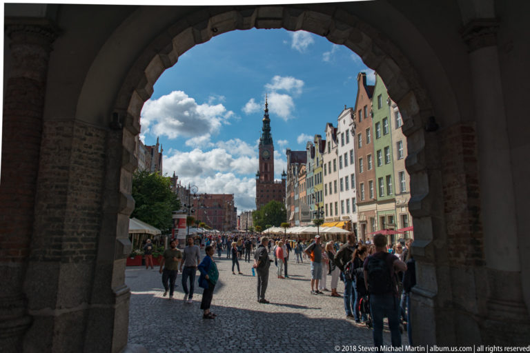 View through a shadowed brick arch toward a cobbled pedestrian street, colorful gabled façades, crowds, and a distant tower.