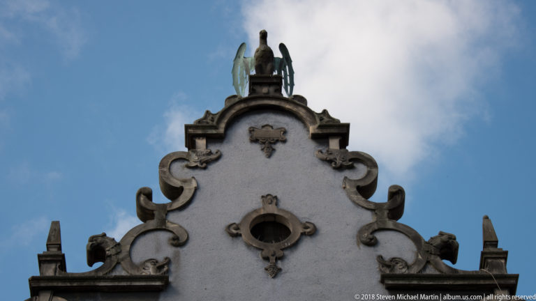 Pale scrolling gable with oval opening, relief ornaments, finials, and a seated winged figure at the summit.