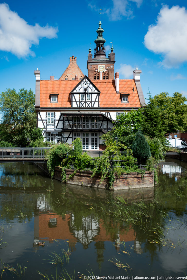 Half-timbered building with red tiled roof reflected in calm canal water amid trees.