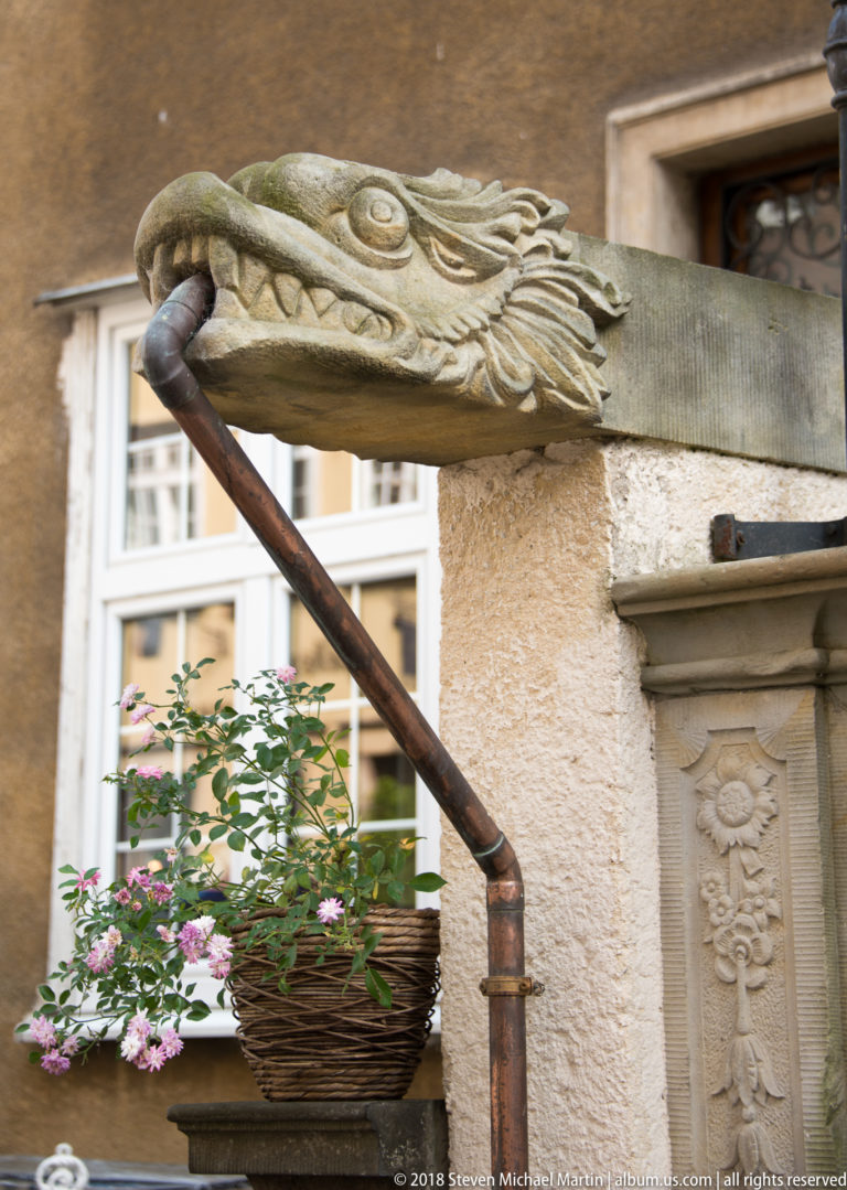 Long metal waterspout ending in a carved dragon-like head above a basket of pink flowers on a historic façade.