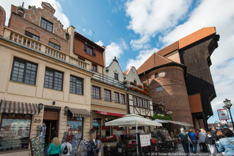 Gdańsk Crane towering behind waterfront cafés, shops, pedestrians, and historic façades under a blue sky.
