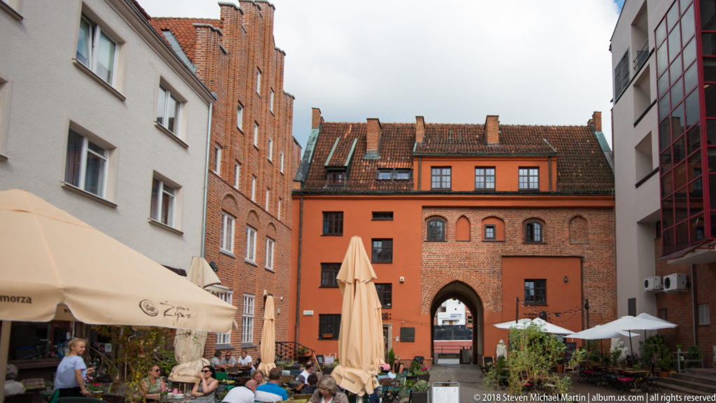 Outdoor café courtyard between brick historic buildings, ending at a pointed-arched gateway beneath a tiled roof.
