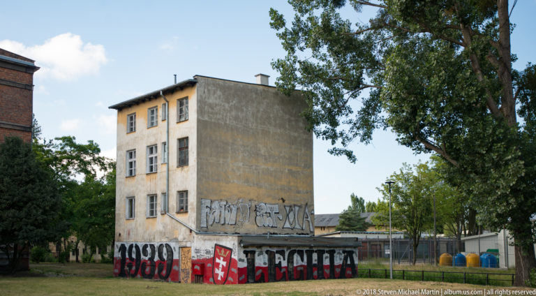 Weathered multi-story building beside grass and trees, with large graffiti covering the lower walls and blank side façade.