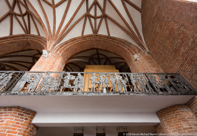 Decorative metal gallery with figurative panels beneath red-brick arches and patterned rib vaults in a church interior.