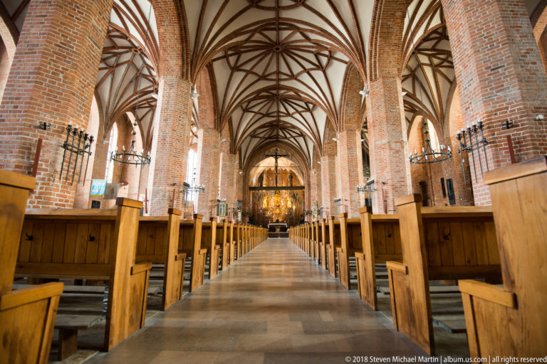 Central aisle of a Brick Gothic church with wooden pews, tall brick piers, chandeliers, and rib vaulting.