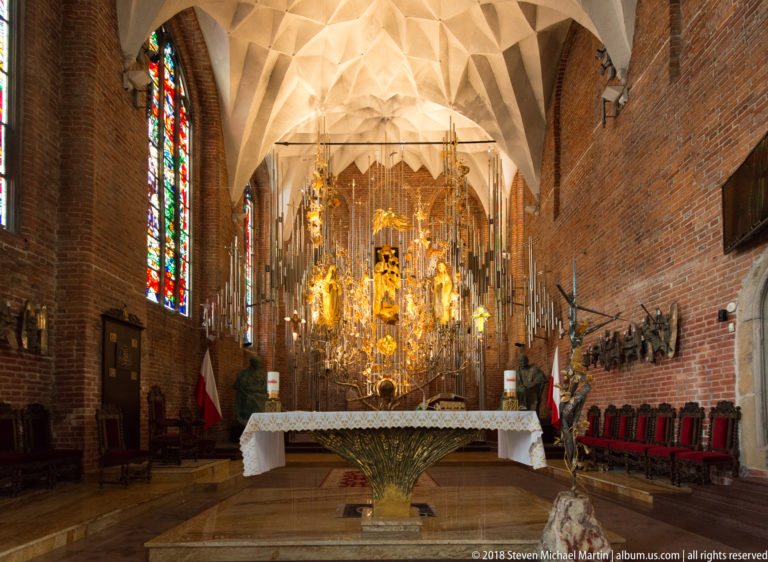 Ornate gilded church altar beneath pale Gothic vaulting, with stained-glass windows and brick walls.