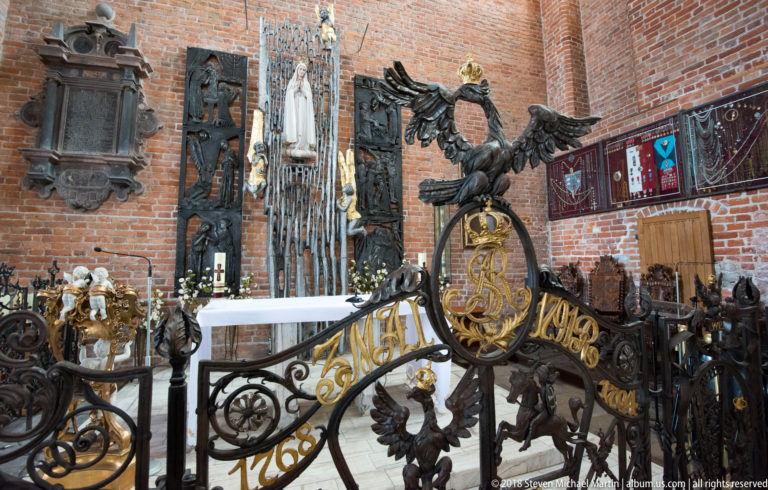 Ornate black-and-gold wrought-iron church screen with eagle crest, monogram, heraldic birds, and dated inscriptions.