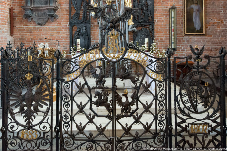 Frontal view of black-and-gold church gate with crowned monogram, heraldic emblems, birds, scrollwork, and gilded dates.