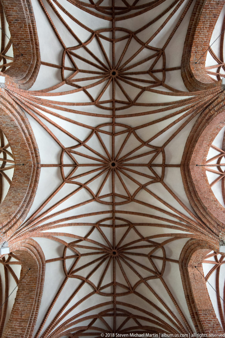 Upward view of pale church vaults crossed by intricate red-brick ribs forming geometric star and polygon patterns.