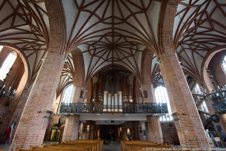 Large pipe organ on an elevated gallery between brick piers beneath intricate Gothic rib vaulting.