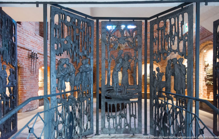 Three tall dark metal panels with cutout human figures, birds, and organic forms inside a brick building.